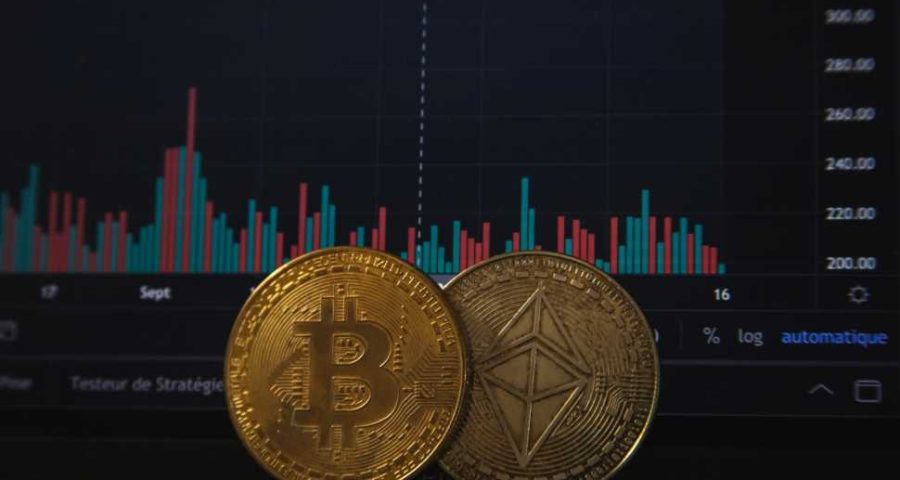 Foto de criptomoedas, Bitcoin e Ether. Ao fundo, um gráfico de oscilações de ações